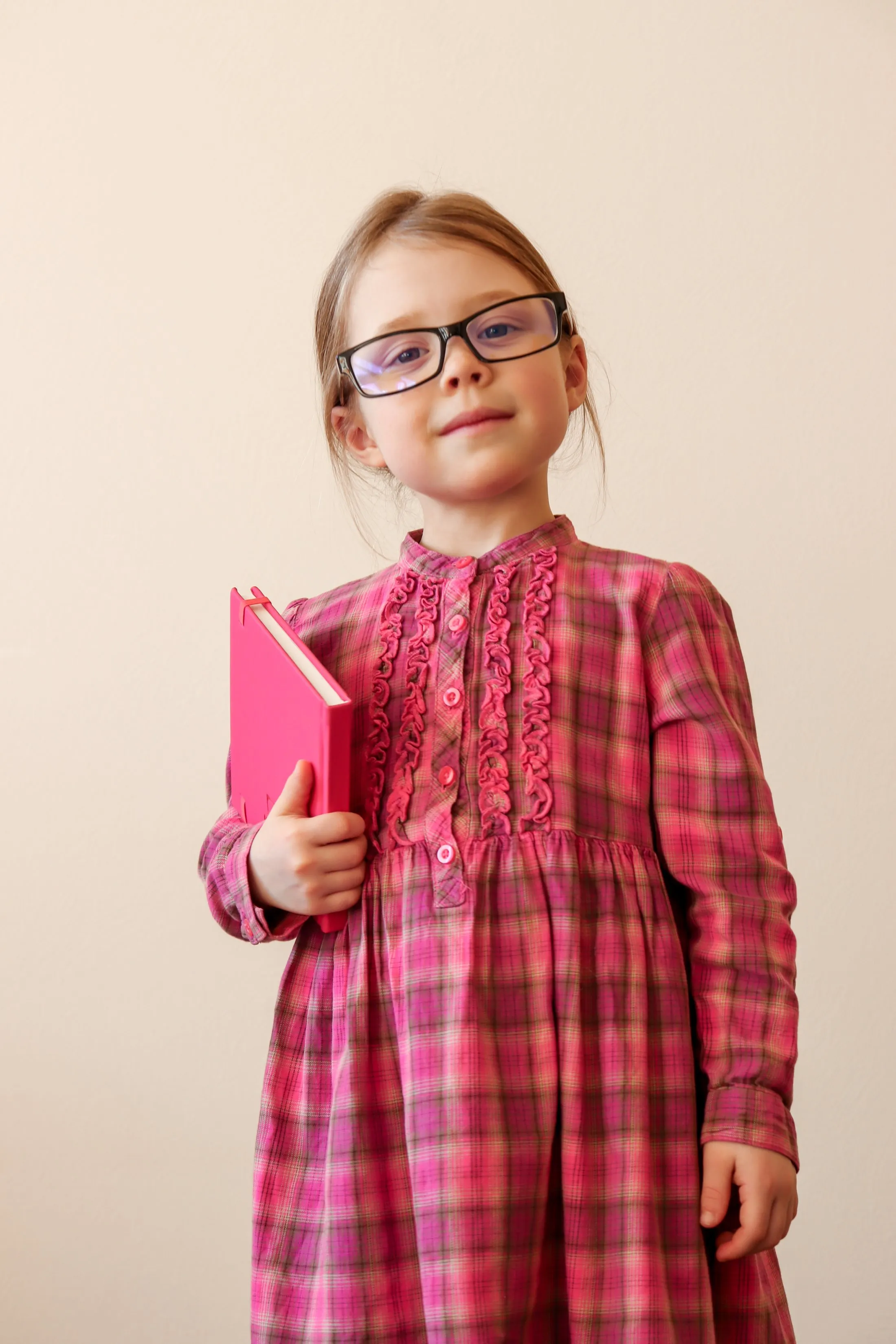 Young girl wearing glasses and pink plaid dress holding a bright pink book
