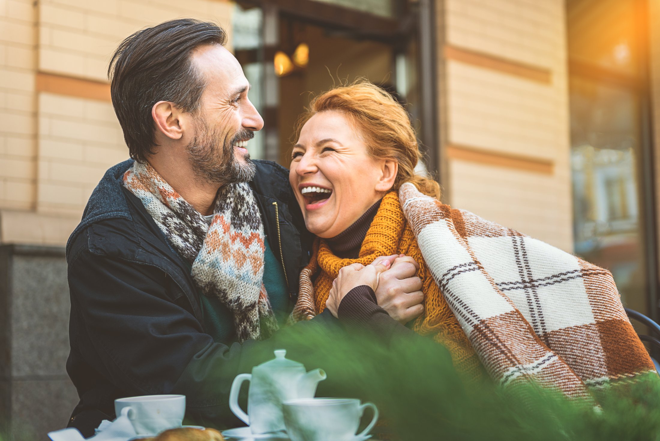 Smiling couple wearing scarves enjoying outdoor tea together in urban setting
