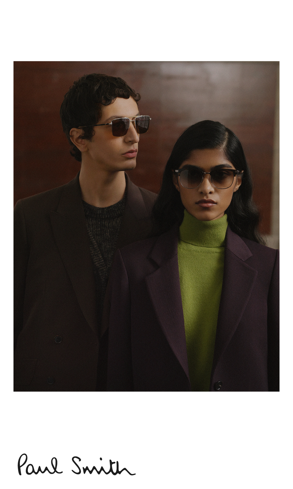 Two people wearing sunglasses and dark blazers pose against a brown background.