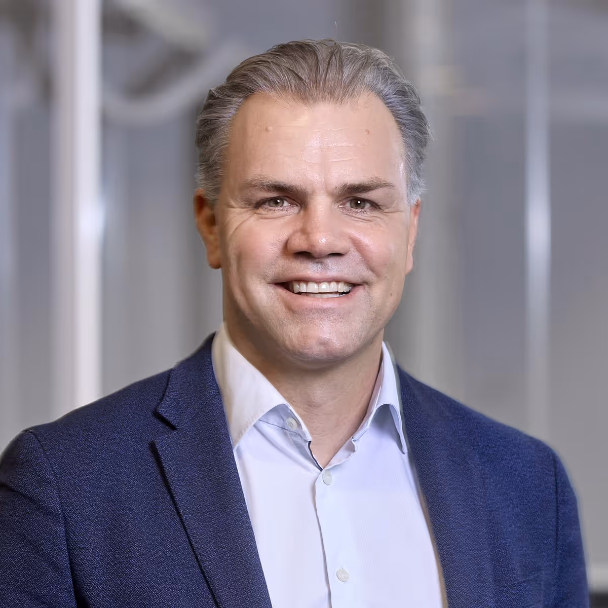 Porträttfoto av en leende man, Ola Klingenborg, med kort grått hår, klädd i blå kavaj och ljus skjorta, sittande inomhus mot en mjukt suddig bakgrund.