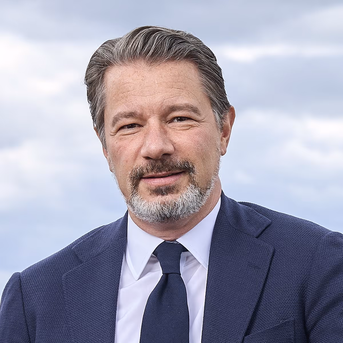 Porträttfoto av en man med kort grått hår och skägg, klädd i kostym och slips, som ser in i kameran mot en ljus, suddig bakgrund.