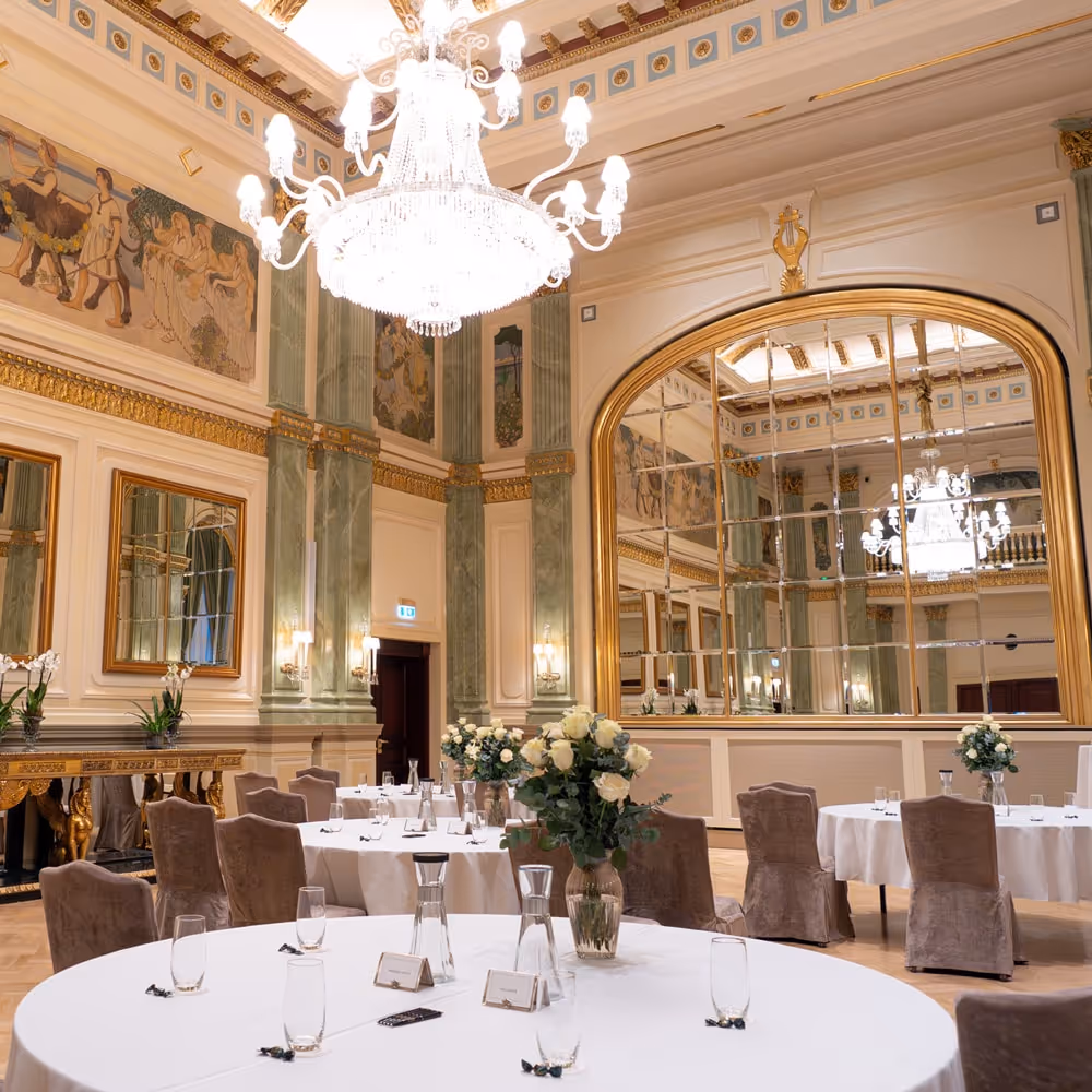 Foto av en elegant sal med högt i tak, kristallkrona och stora speglar med guldram. Runda bord med vita dukar, glas och blomsterarrangemang står utplacerade i rummet.
