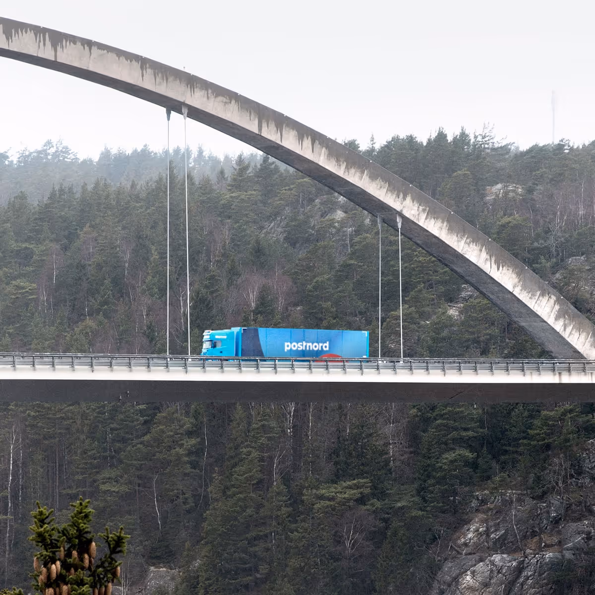 Foto av en lastbil med Postnord-logotyp som kör över en hög bro med bågformad konstruktion, omgiven av skog och berg i bakgrunden.