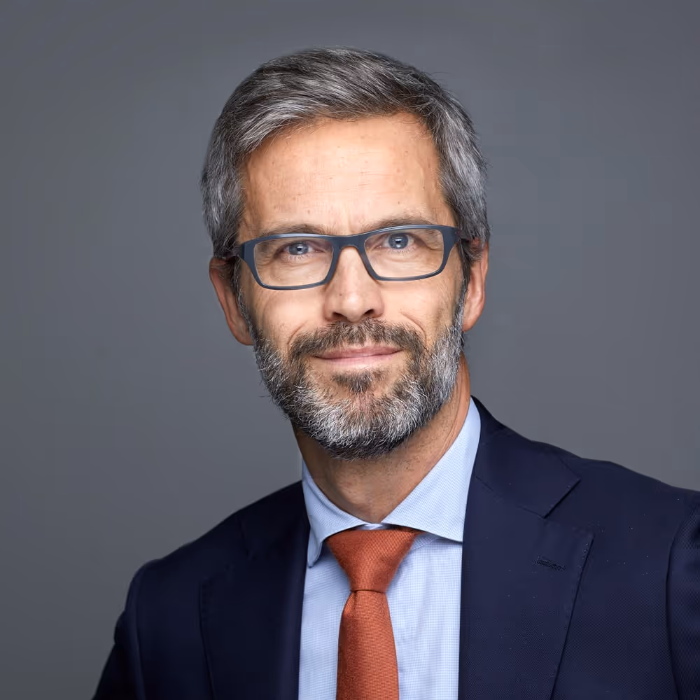 Porträttfoto av en man med glasögon, Jens Ebbe Rasmussen, kort grått hår och skägg, klädd i kostym, ljus skjorta och röd slips, mot en neutral grå bakgrund.