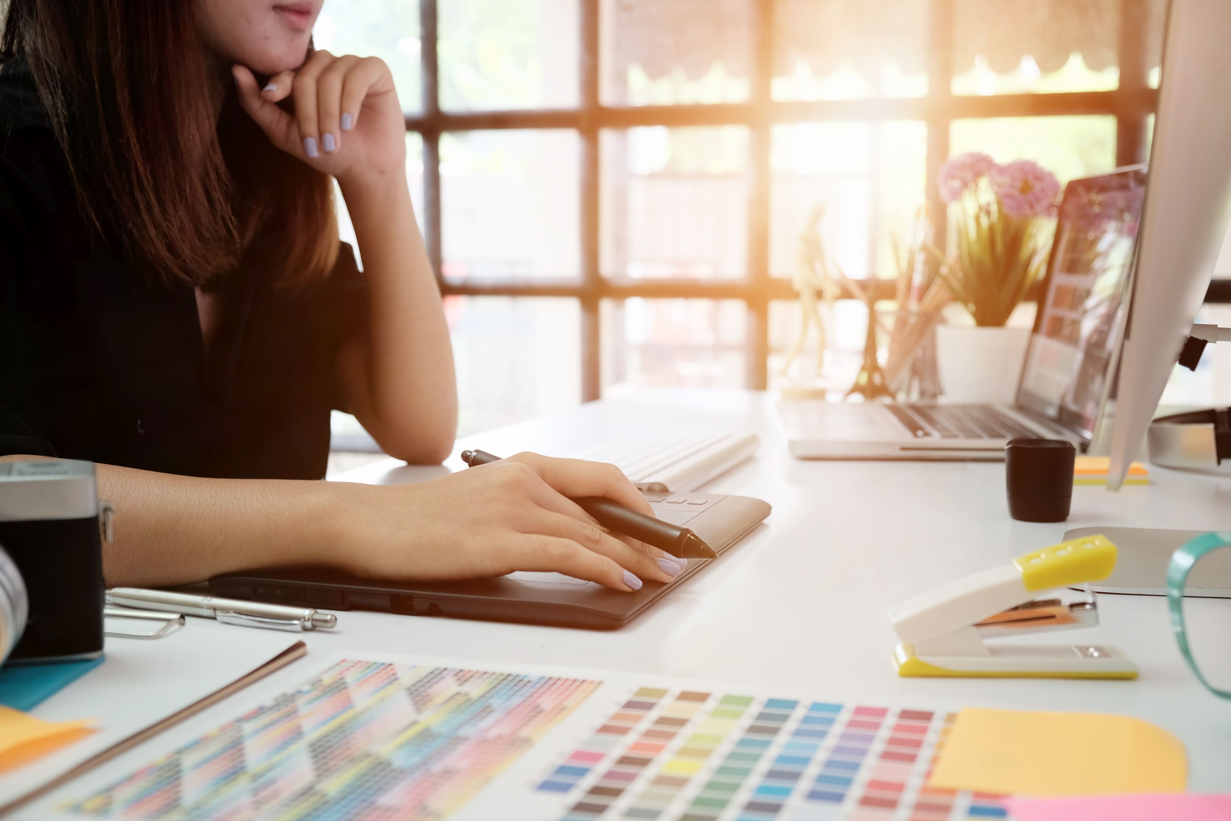Person using a stylus pen on a graphic tablet at a bright desk with a color chart, laptop, and office supplies.