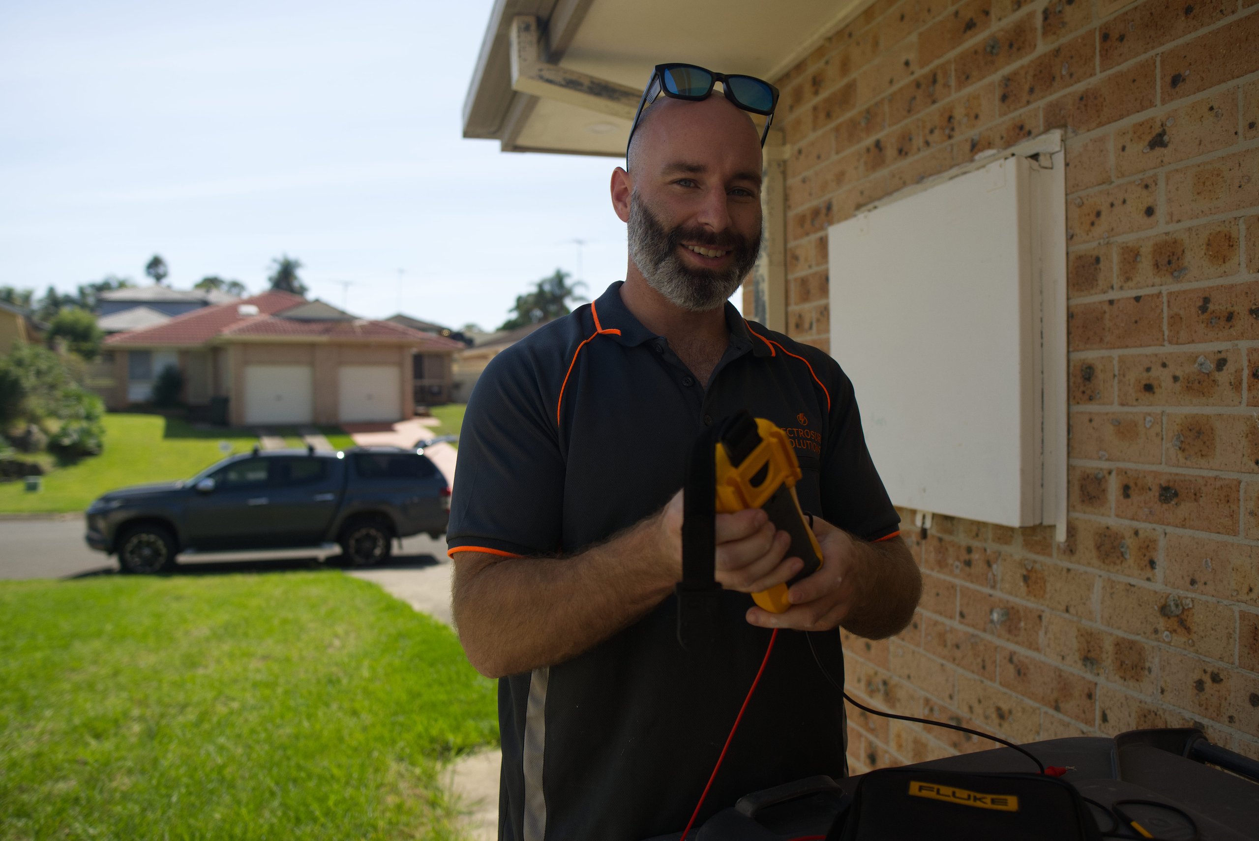 Smiling technician holding a yellow electrical testing device outside a brick house with outdoor equipment nearby.