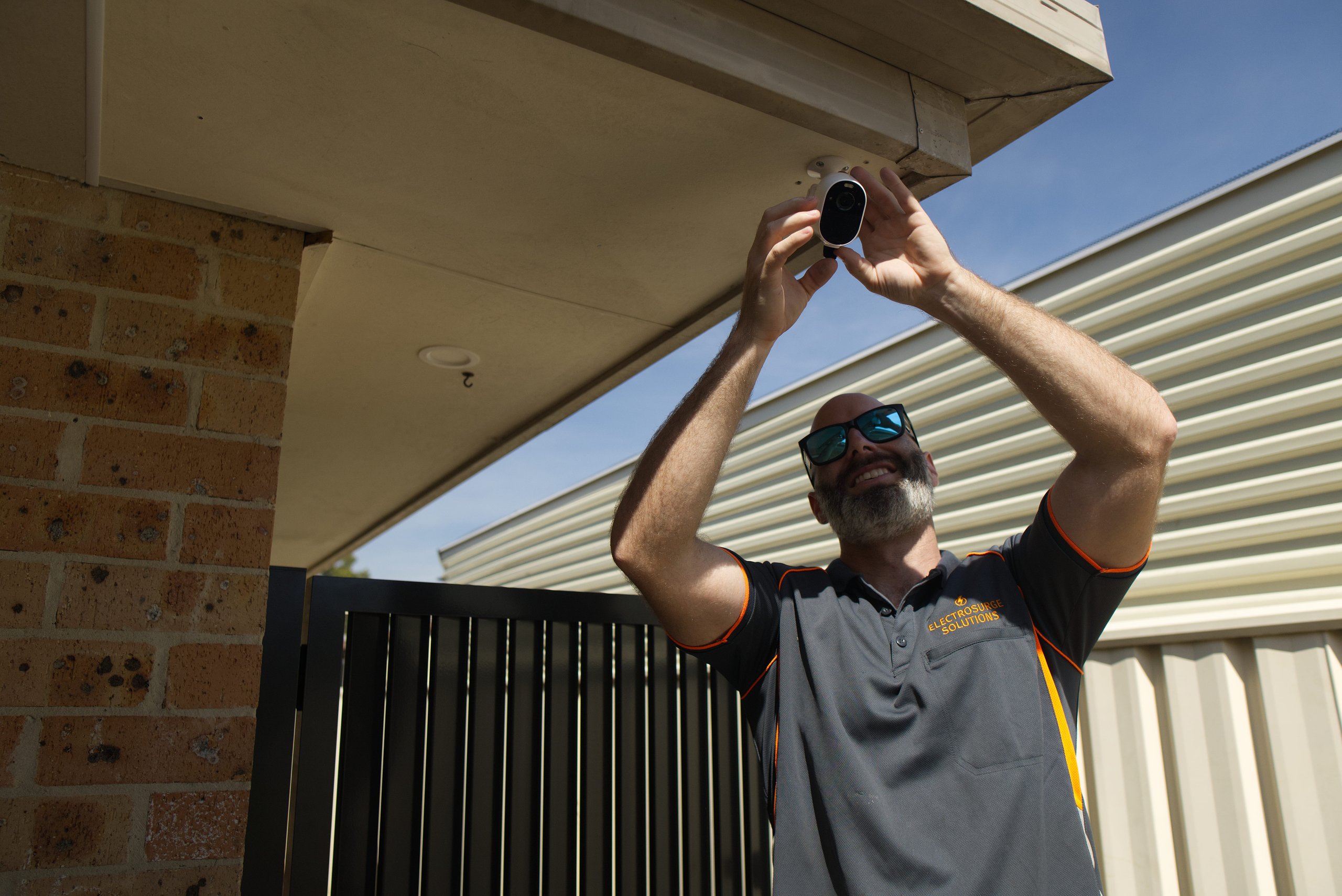 Bearded man in sunglasses installing a security camera under the eave of a house.