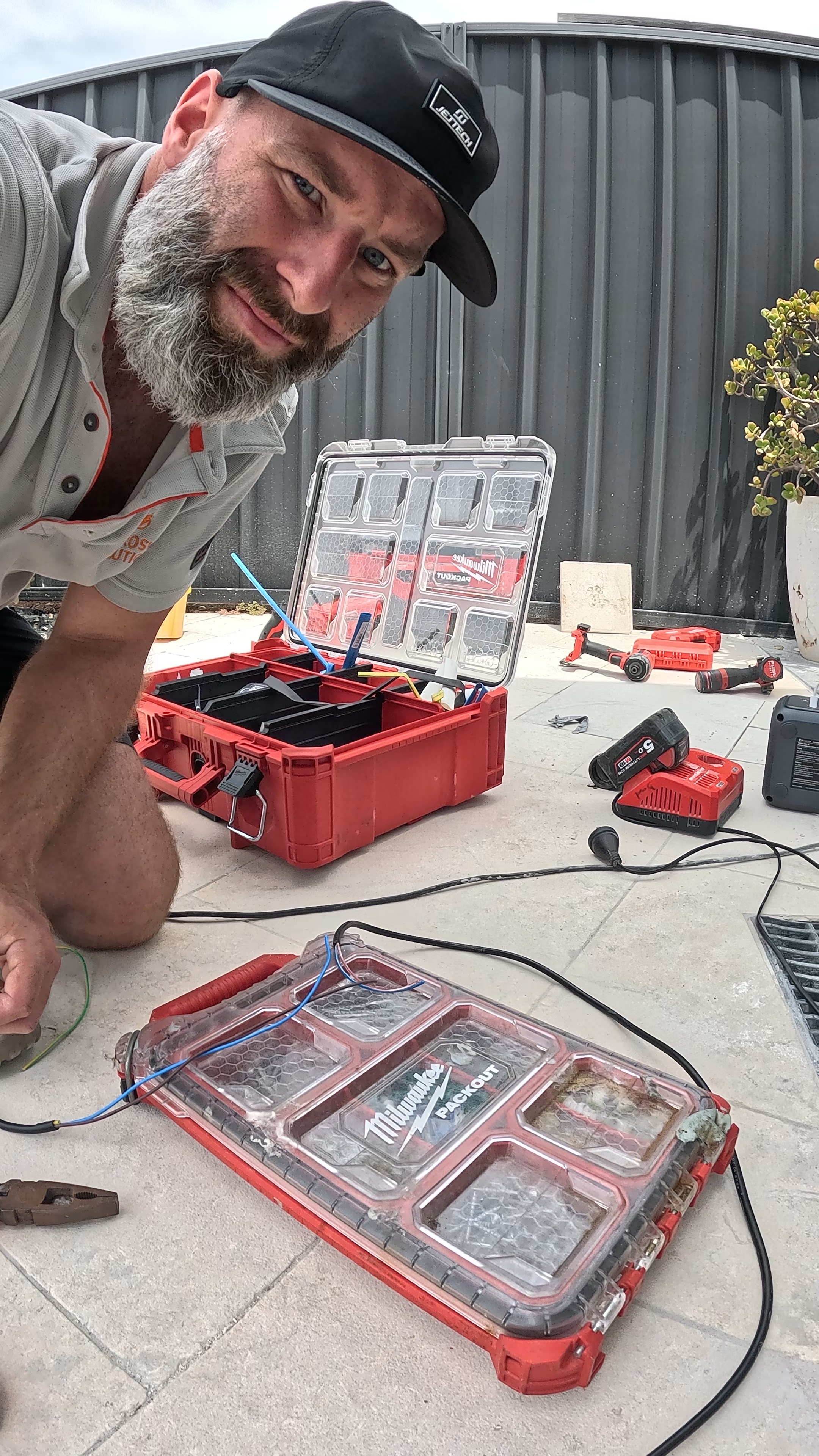 Electrician wearing a black cap kneeling on a tiled floor with red Milwaukee Packout toolboxes and power tools scattered around.