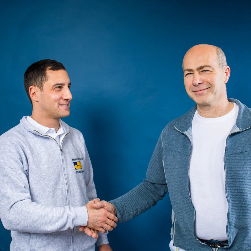 Zwei Männer (Der ehem. Inhaber Roland Schmid und der neue Inhaber Pierre Moll) geben sich vor blauem Hintergrund die Hand, einer trägt eine graue Jacke mit einem Firmenlogo.