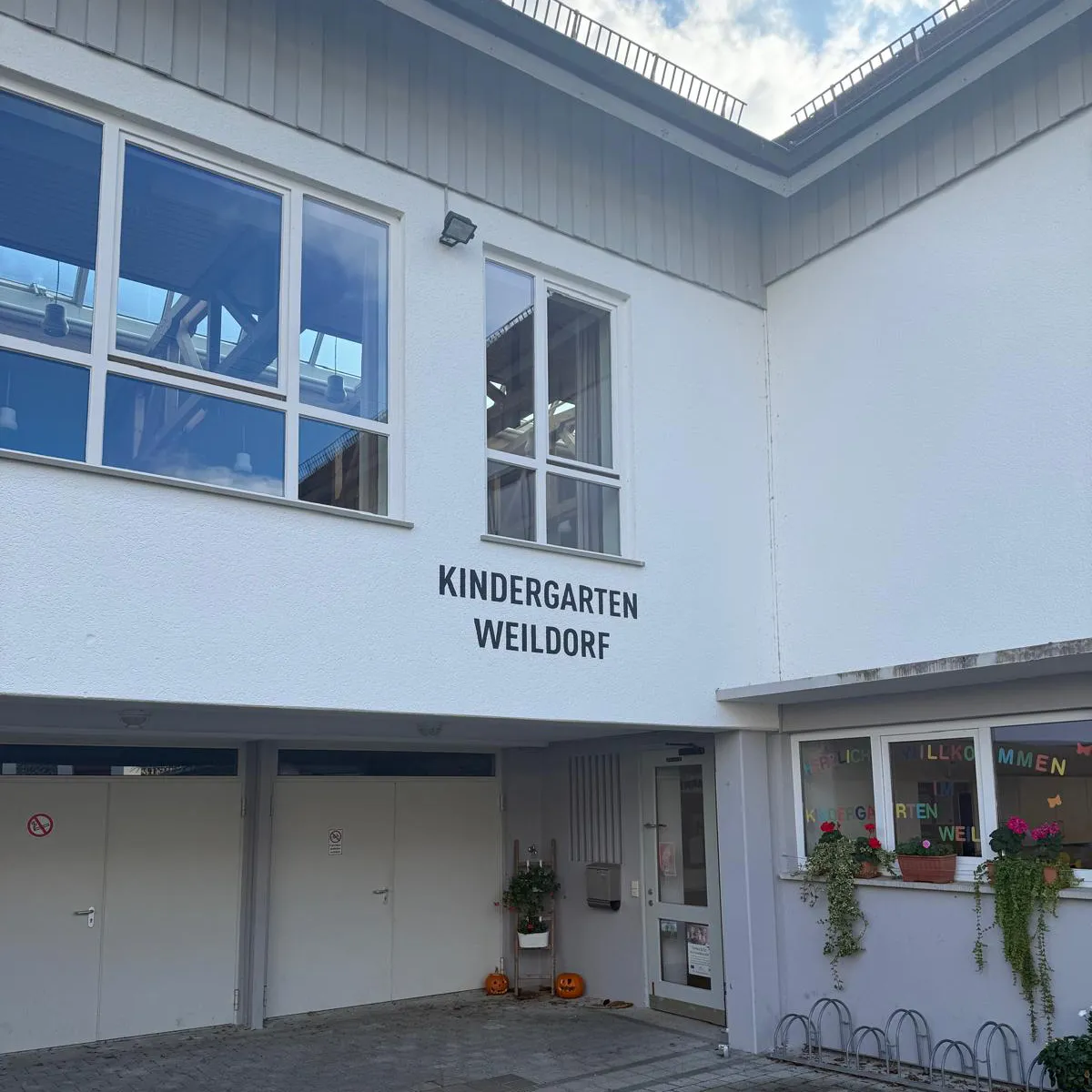 Außenansicht des Kindergarten Weildorf mit weißen Wänden, Fenstern und dekorativen Pflanzen in Blumentöpfen.