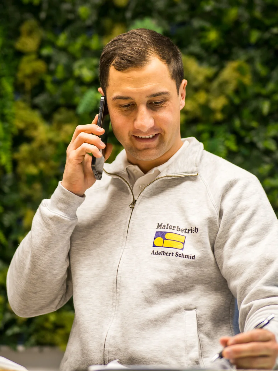 Der Inahber Pierre in grauem Pullover mit Schriftzug 'Malerbetrieb Adelbert Schmid' telefoniert und macht sich Notizen.