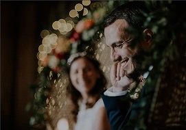 Smiling groom with hand near face and blurred bride in background during wedding ceremony decorated with flowers and lights.