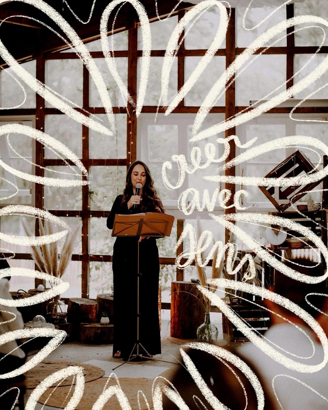 Woman in black dress speaking into a microphone, standing at a music stand in a rustic room with wooden beams and large windows.