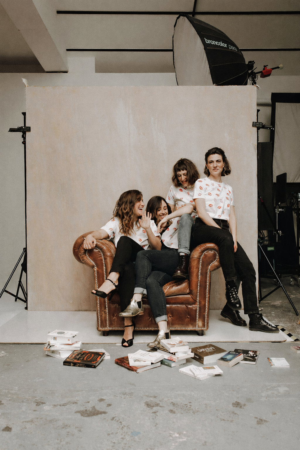Four women wearing white shirts with red lip prints sitting and posing on and around a brown leather armchair in a photography studio with scattered books on the floor.