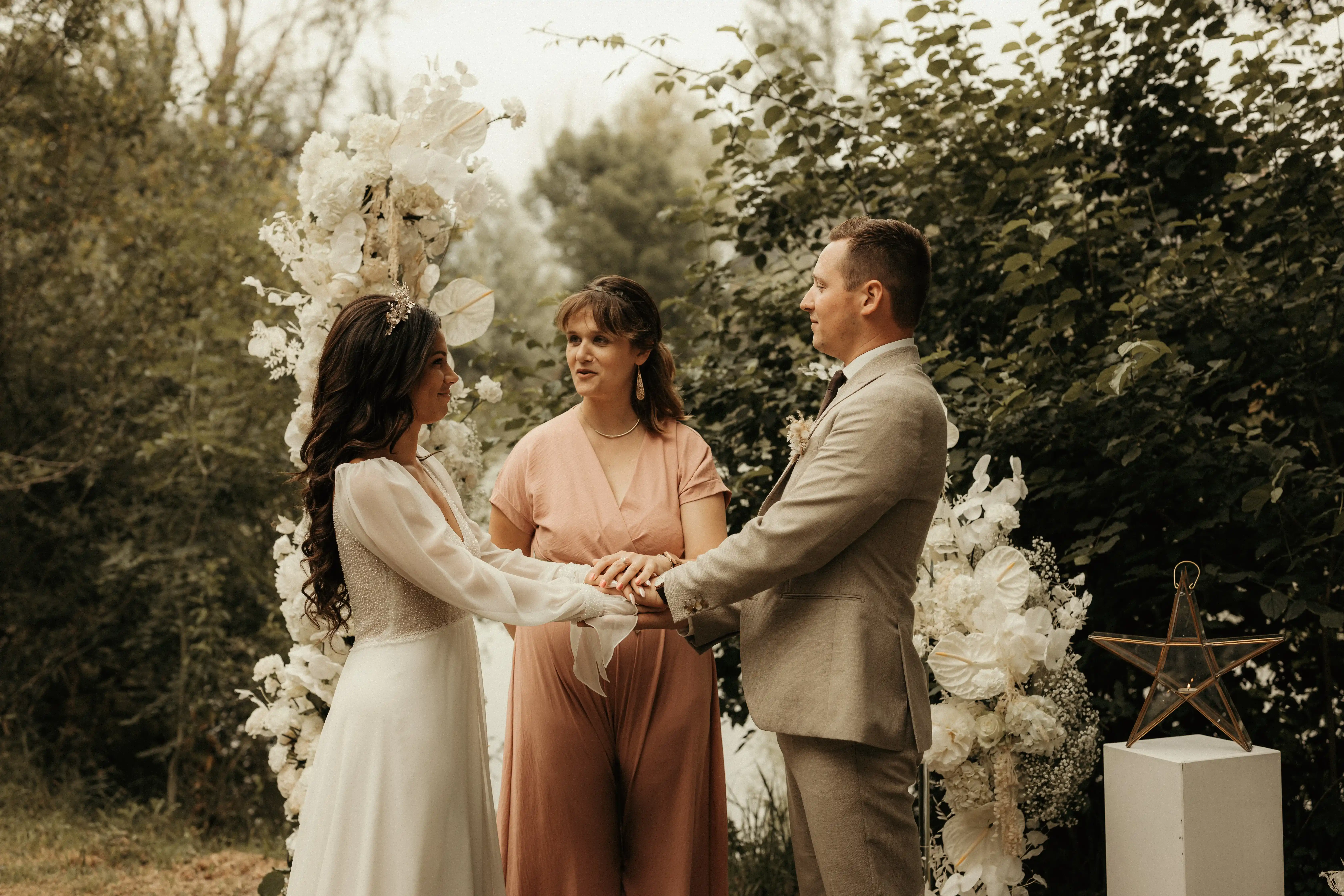 Bride and groom holding hands with officiant during outdoor wedding ceremony with floral decorations.