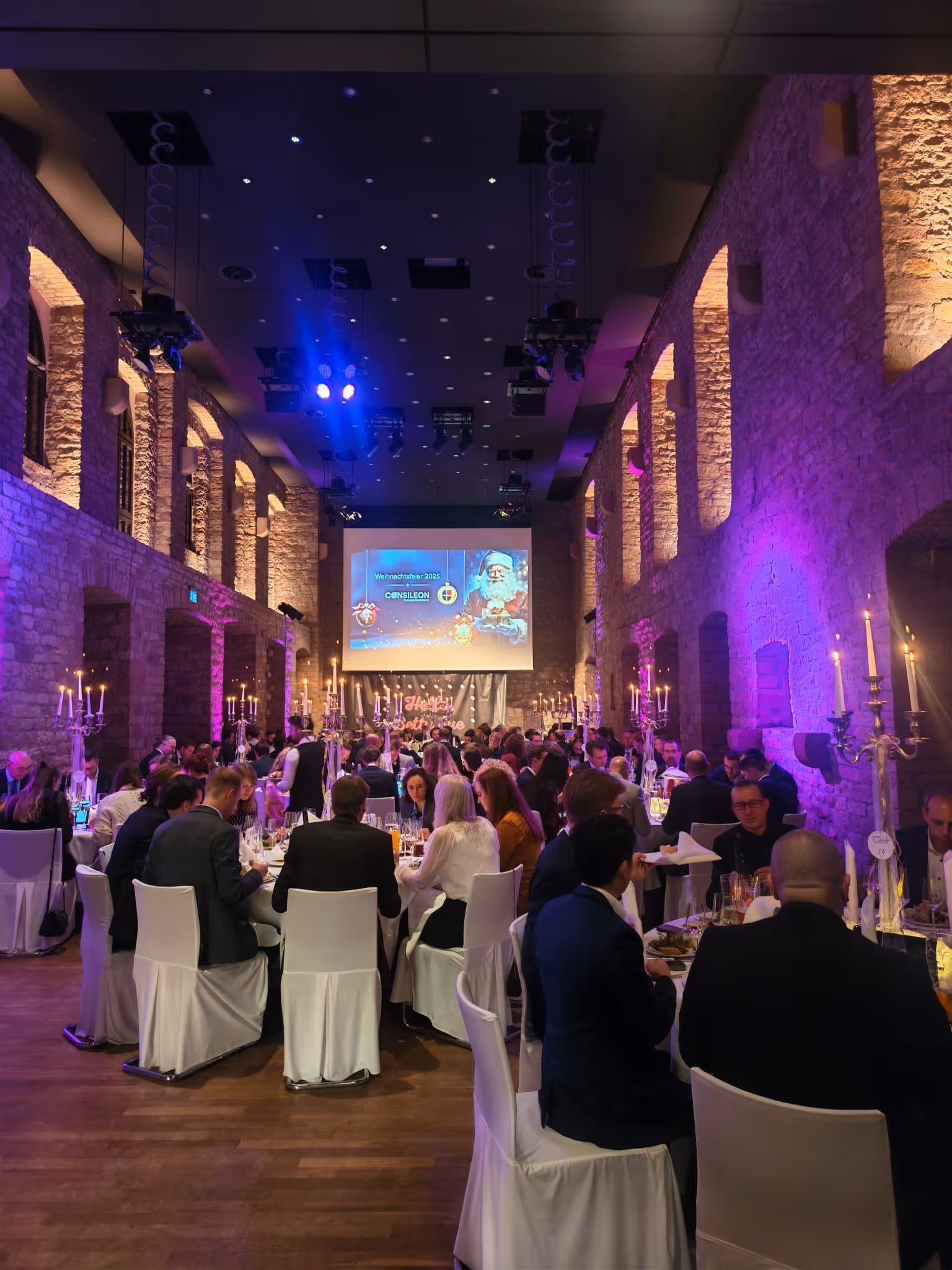 Festlich gekleidete Menschen sitzen in einem großen Saal mit Steinwänden und Kronleuchtern an gedeckten Tischen bei einer Weihnachtsfeier.