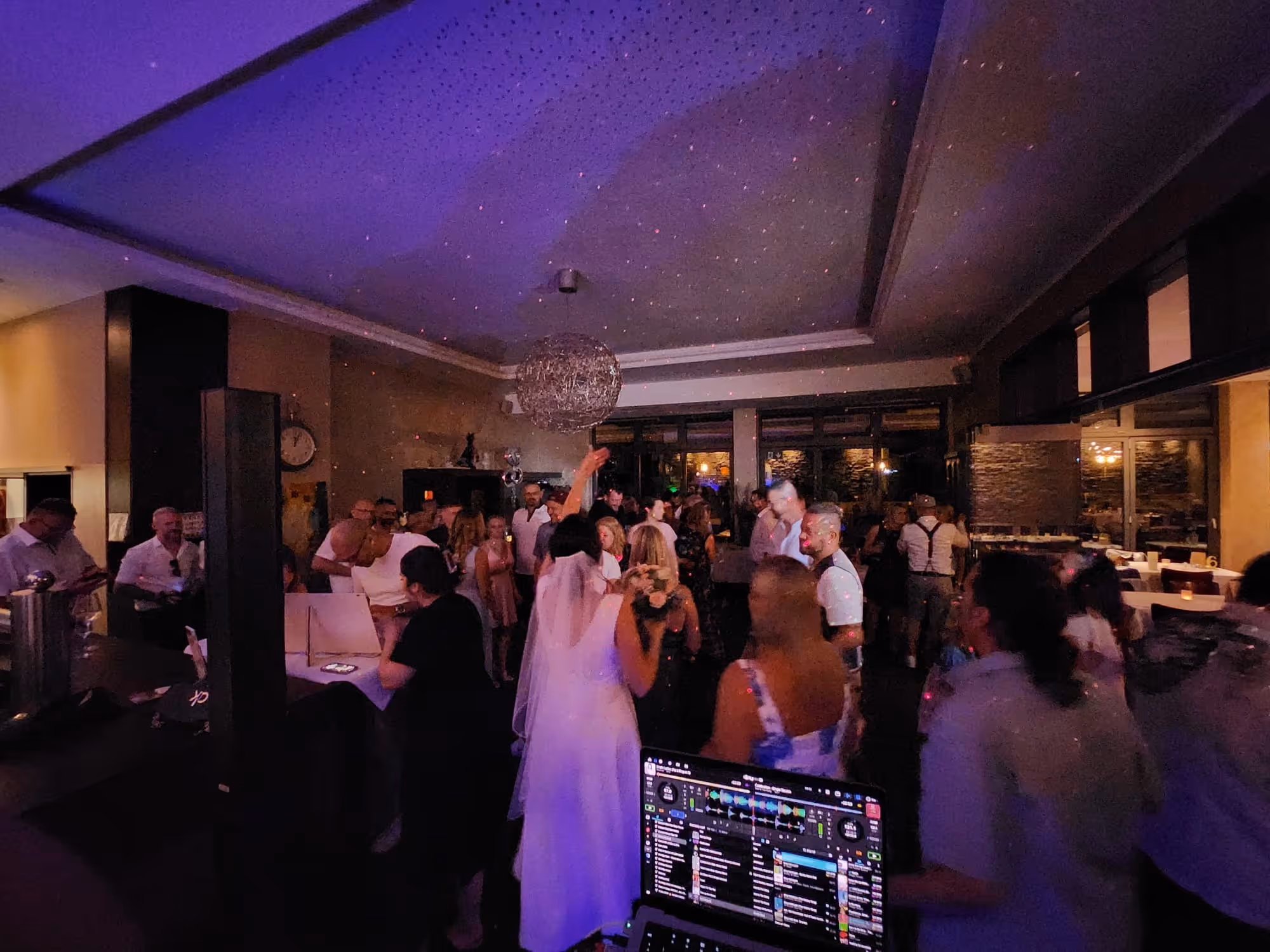 Menschen tanzen und feiern in einem schummrig beleuchteten Raum mit DJ-Equipment im Vordergrund.