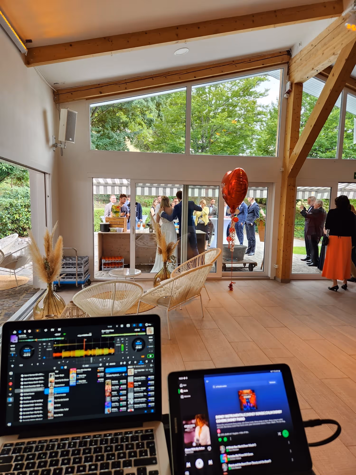 Innenraum mit Holzrahmen und großen Fenstern, zwei offenen Laptops vorne, im Hintergrund Menschen bei einer Feier mit rotem Herzluftballon.