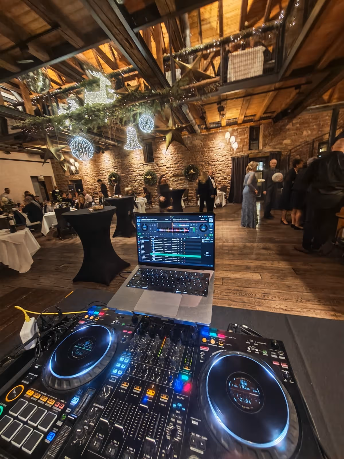 DJ-Setup mit beleuchteten Controllern und Laptop auf einem Tisch vor einer festlich dekorierten Veranstaltung in einem rustikalen Saal mit Holzbalken und Weihnachtsbeleuchtung.