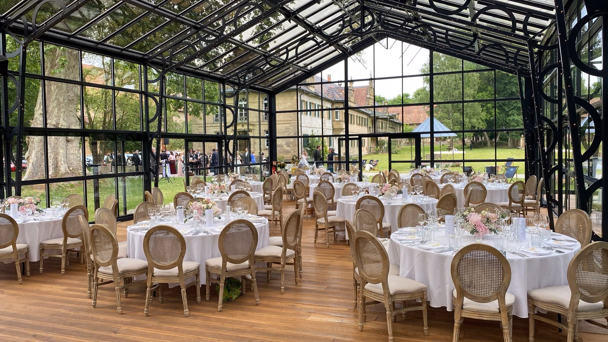 Eleganter Veranstaltungsraum mit runden Tischen, weiß gedeckt und mit Blumenarrangements, in einem Glasgewächshaus mit Blick auf grünen Garten und Gebäude.
