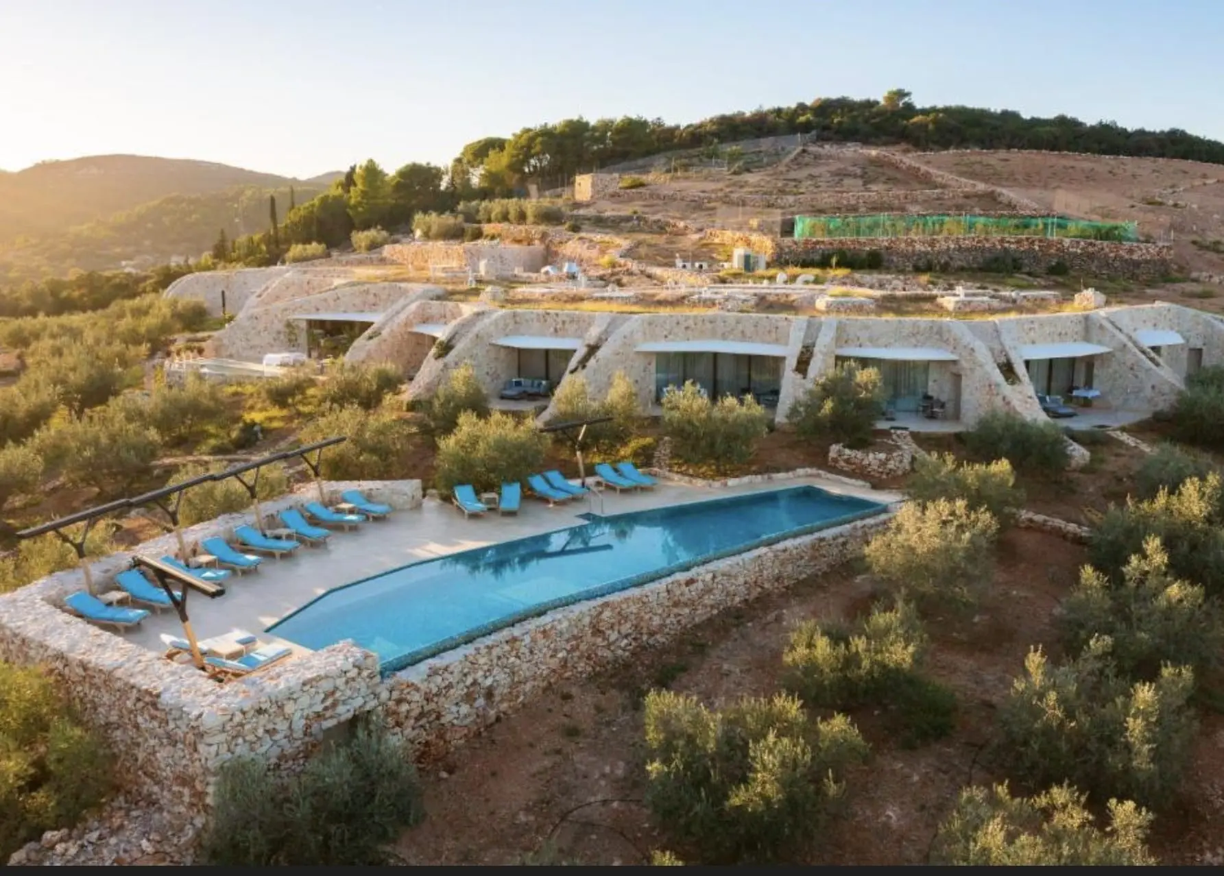 Moderne Steinvillen mit privaten Terrassen und einem Pool mit blauen Liegestühlen in einer hügeligen, bewaldeten Landschaft.