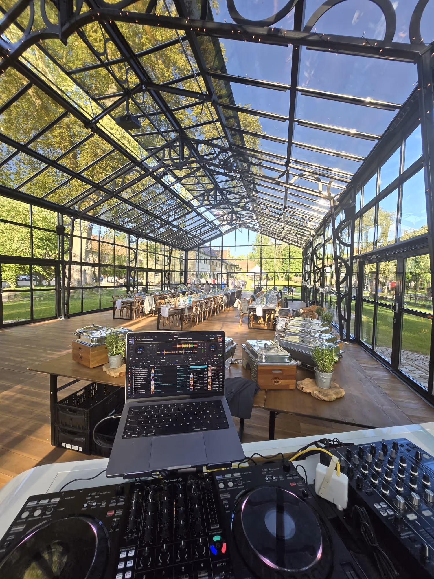 DJ-Mischpult und Laptop in einem Glasgewächshaus mit eingedeckten Tischen für eine Veranstaltung im Freien.