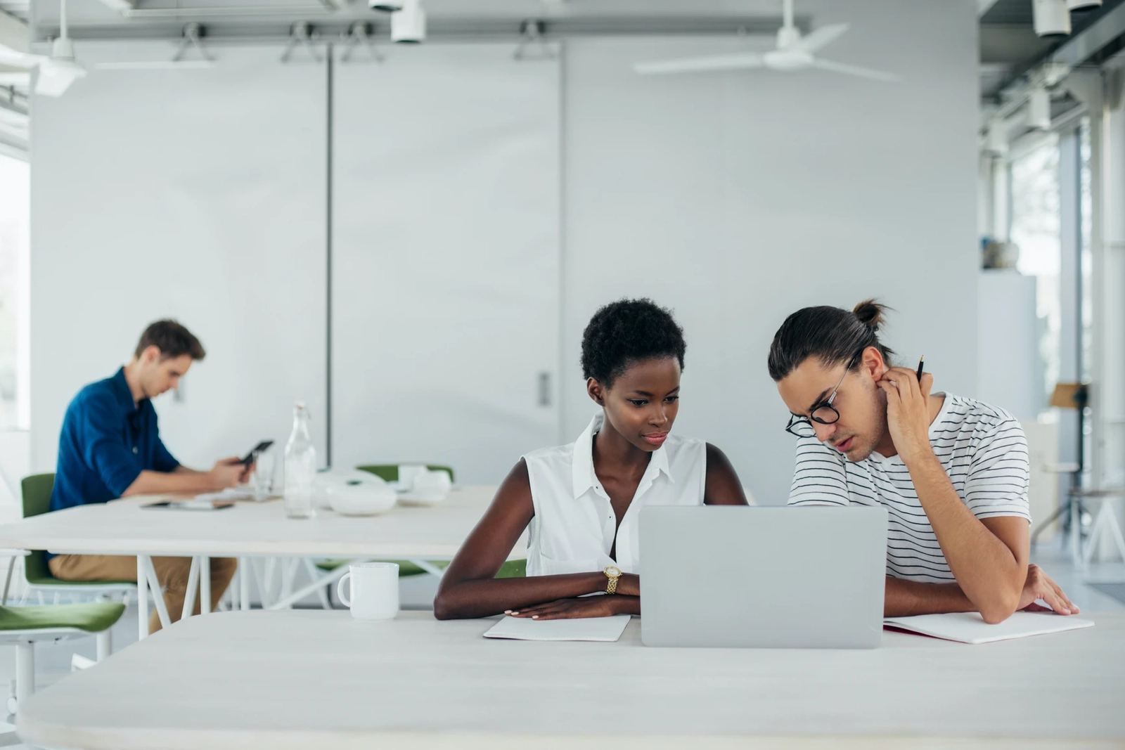 Two young professionals collaborating over a laptop in a bright modern office with a third person working in the background.