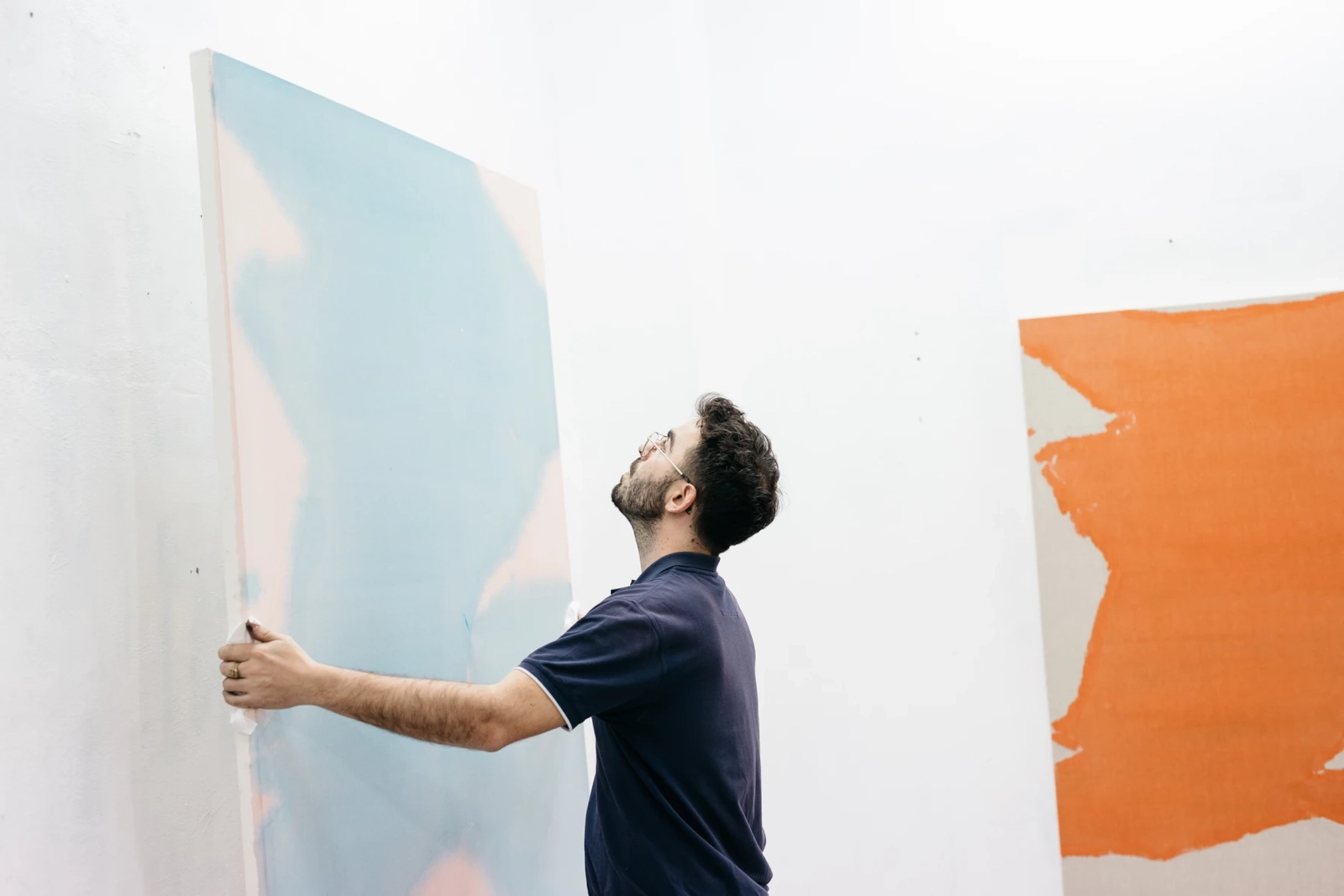 Man with glasses holding and examining a large abstract painting with blue and pink shades in a white gallery space.