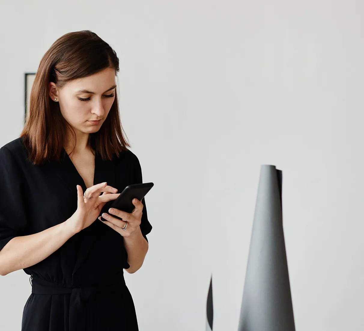 Woman in black dress using a smartphone while standing next to a modern gray metal sculpture on a white pedestal.