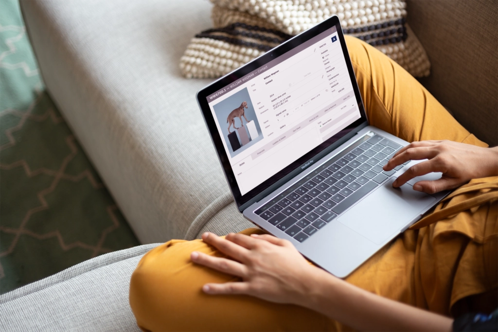 Person sitting on a sofa with mustard yellow pants using a MacBook Pro showing a contact information webpage with a dog image.