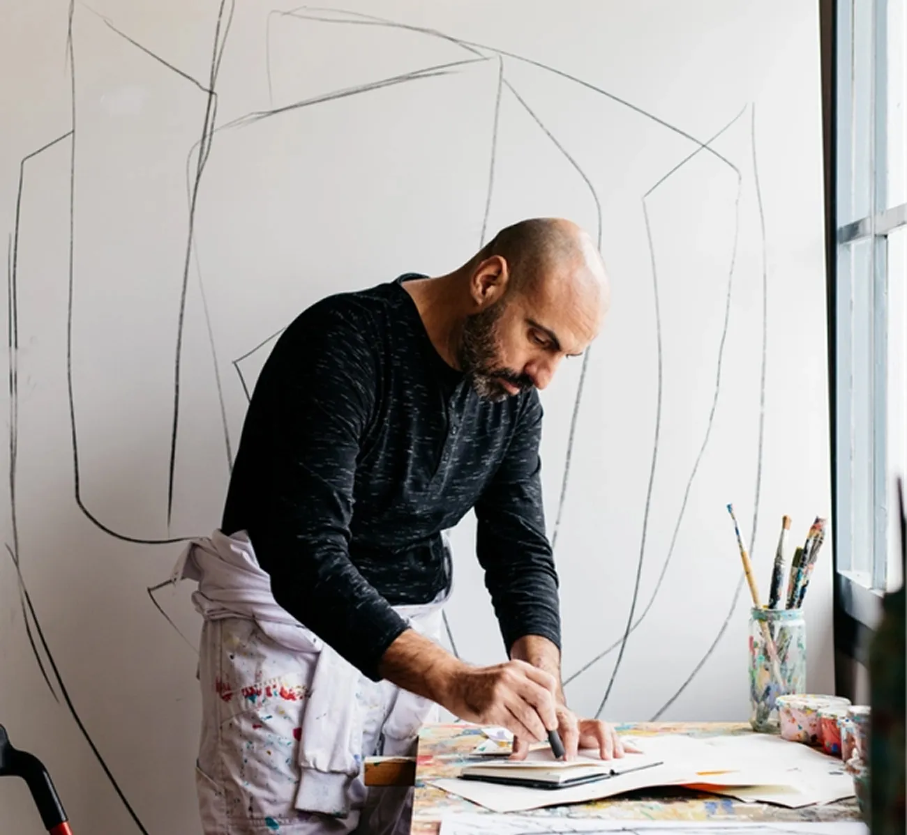 Man with a bald head and beard drawing on paper at a cluttered table with paint brushes in a jar beside him.
