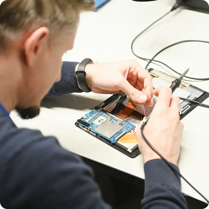 Man repairing electronic circuit board on a tablet with a soldering iron at a workstation.