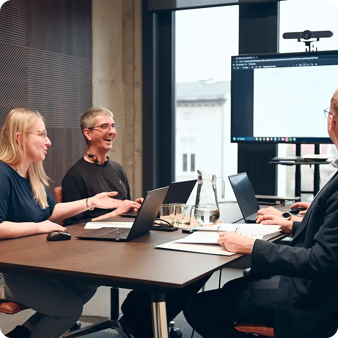 Three colleagues having a meeting around a table with laptops and documents, discussing in a modern office setting with a screen in the background.