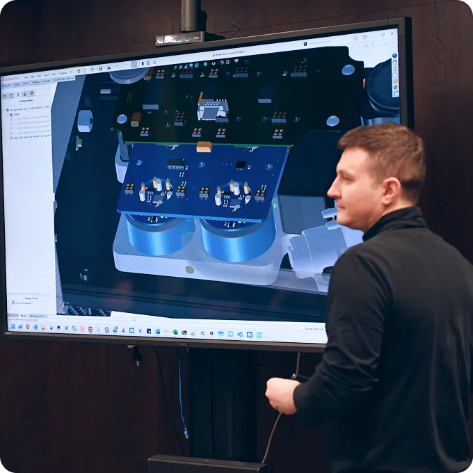 Man standing next to a large screen displaying a 3D CAD design of electronic circuit boards and components.