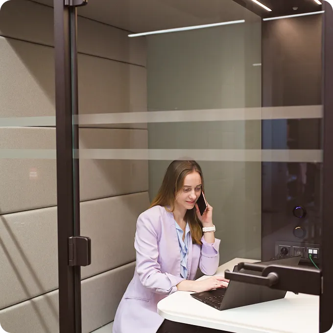 Young woman in a purple blazer sitting in a small office booth, talking on a smartphone and working on a laptop.