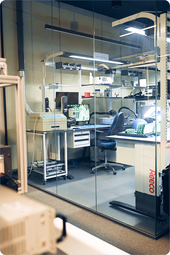 Modern electronics lab with computer monitors, circuit boards, lab equipment, and office chairs behind glass walls.