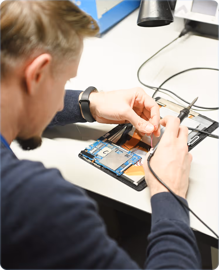 Technician with a smartwatch repairing a circuit board on an electronic device at a workstation.