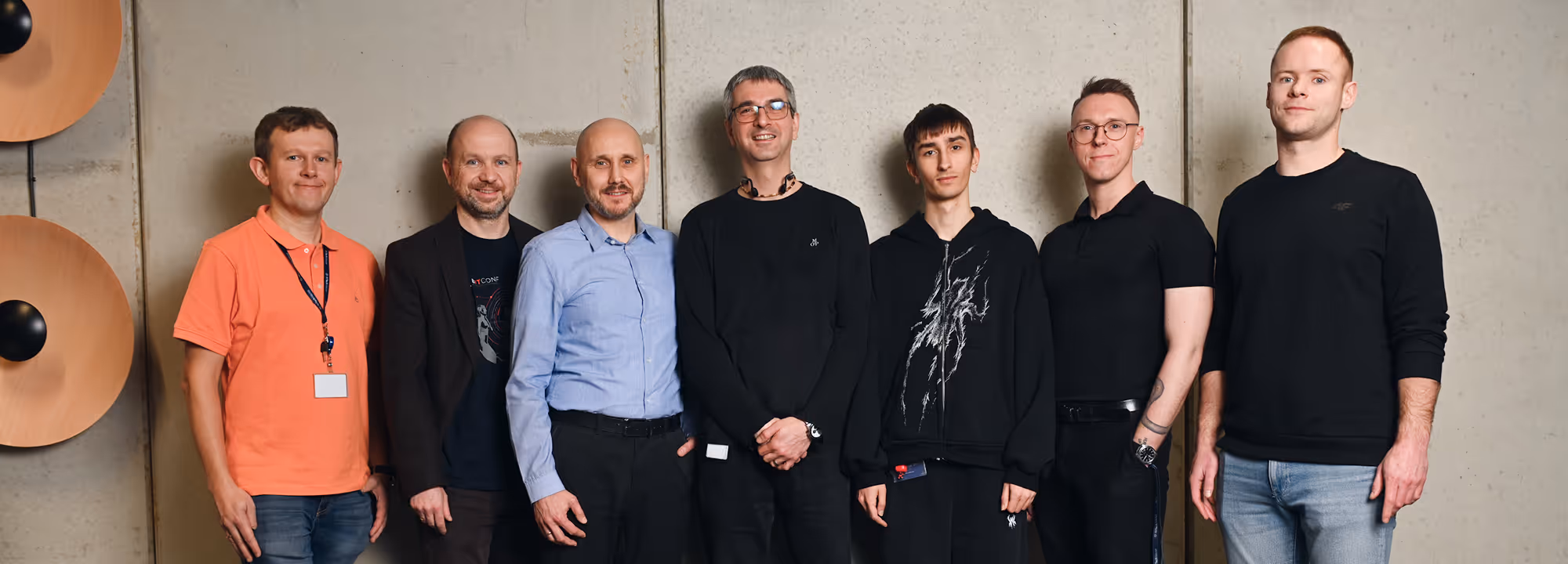 Group of seven men standing in a row against a concrete wall, all looking at the camera.