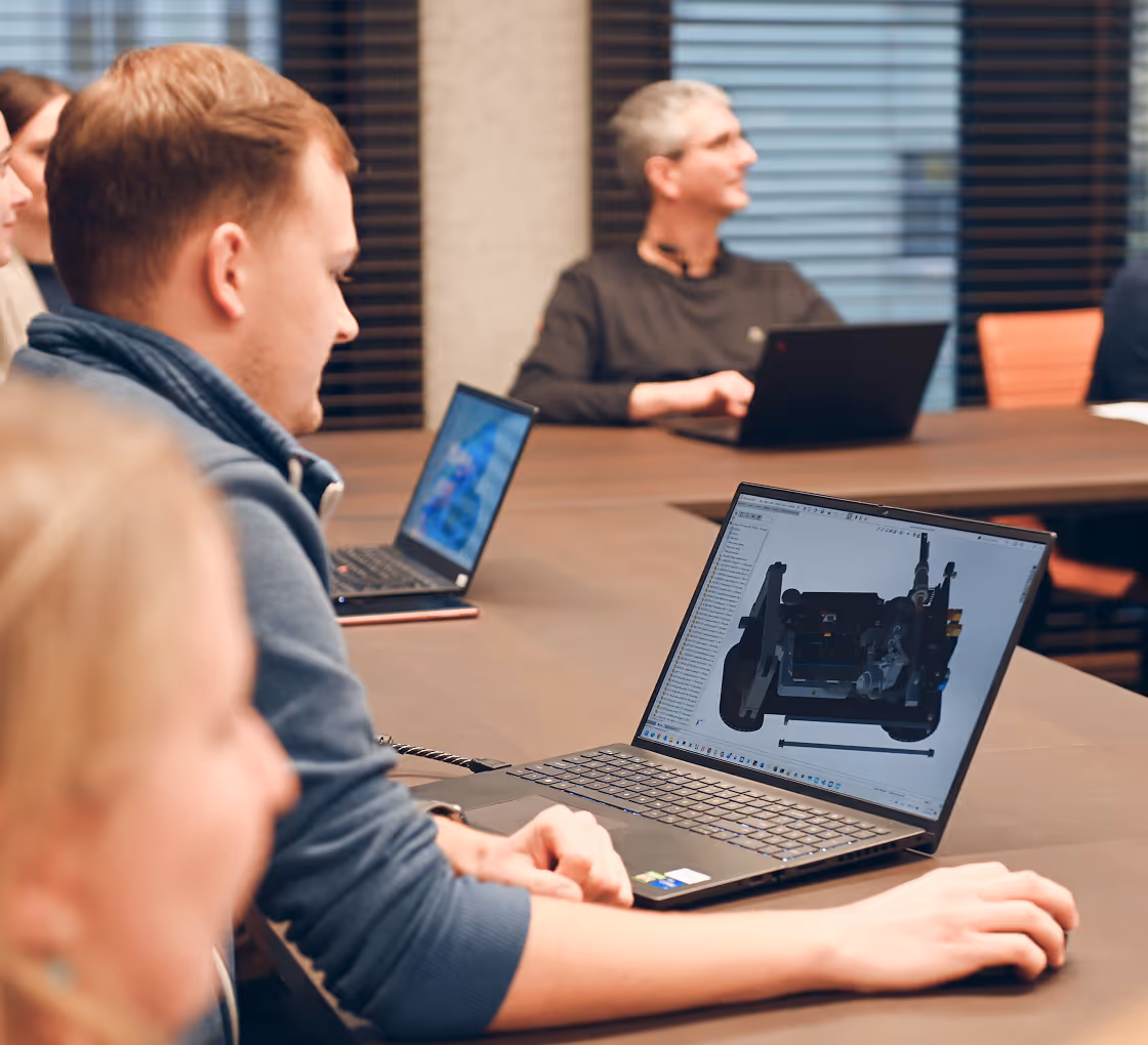 Young man working on a 3D mechanical design on a laptop during a meeting with colleagues at a conference table.
