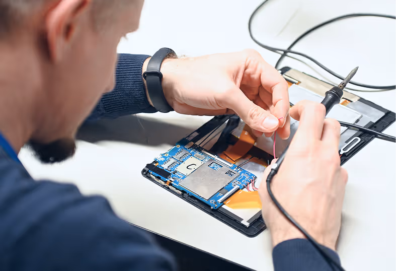 Technician repairing an electronic device circuit board with a soldering iron on a white surface.