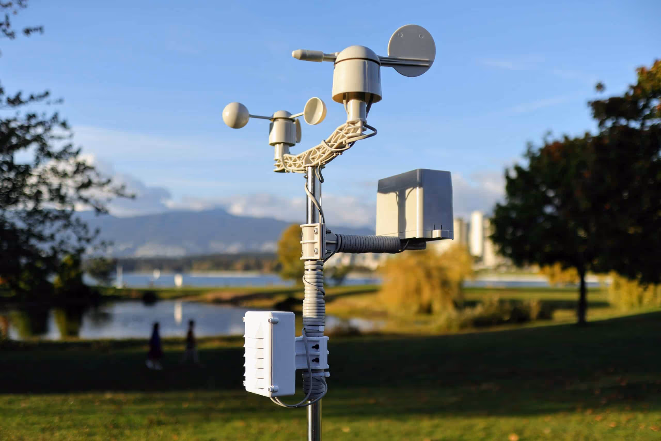 Close-up of an outdoor weather and environmental sensor station equipped with anemometers and other measurement devices, set against a blurred background of a park, water, and distant mountains.