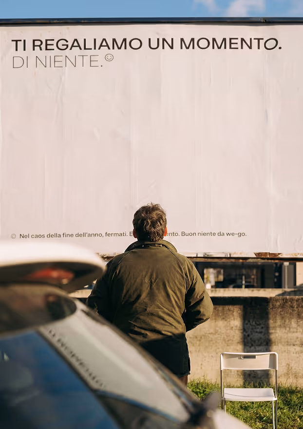 Una persona con giacca verde scuro vista di spalle osserva un grande manifesto con la scritta 'Ti regaliamo un momento. Di niente.' accanto a una sedia bianca e un'auto parcheggiata.