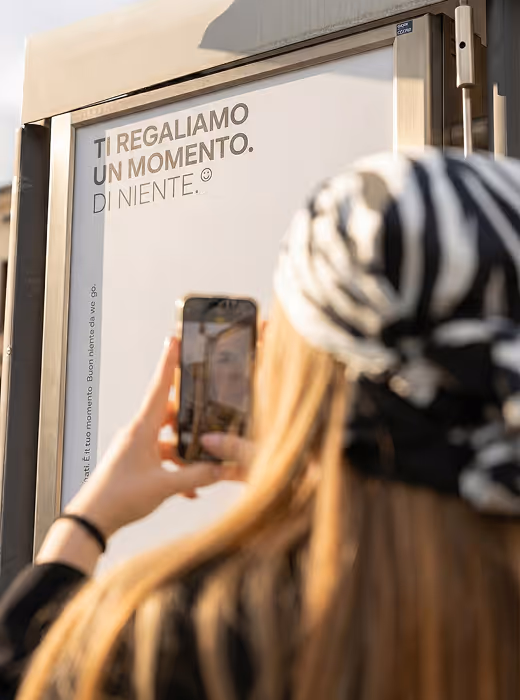 Una donna con berretto a strisce prende una foto con il cellulare di un cartello che dice 'TI REGALIAMO UN MOMENTO. DI NIENTE.'