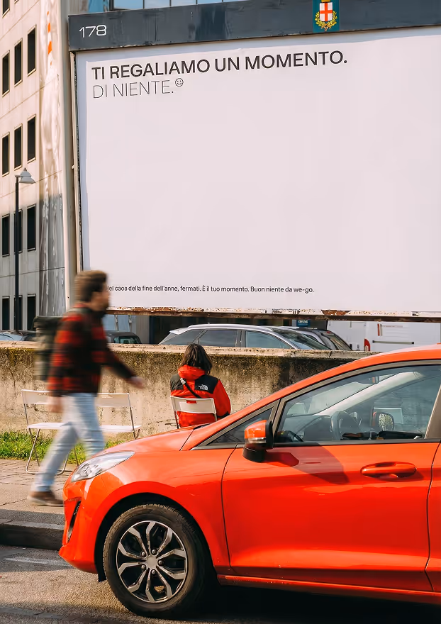 Cartellone pubblicitario con scritto 'Ti regaliamo un momento. Di niente.' e persone davanti a una macchina rossa parcheggiata.