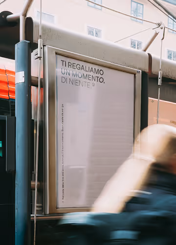 Cartellone pubblicitario urbano con scritto "Ti regaliamo un momento di niente" e persone sfocate in movimento davanti.