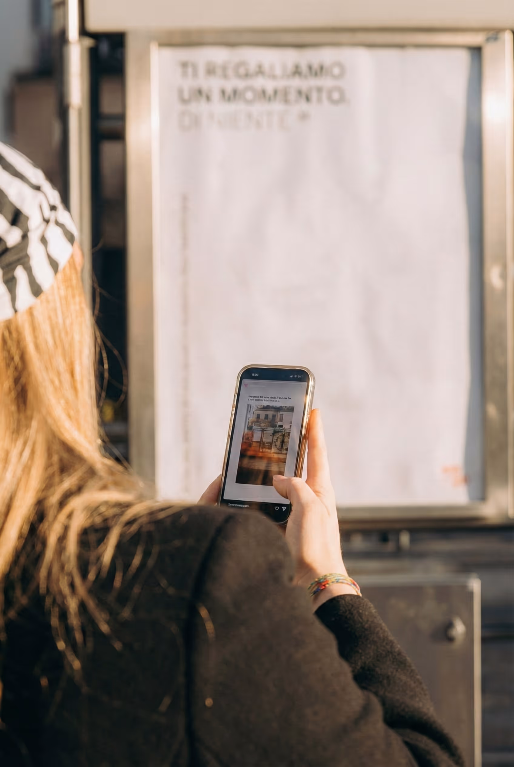 Donna con cappello a righe che guarda il telefono davanti a un cartellone sfocato con testo italiano.