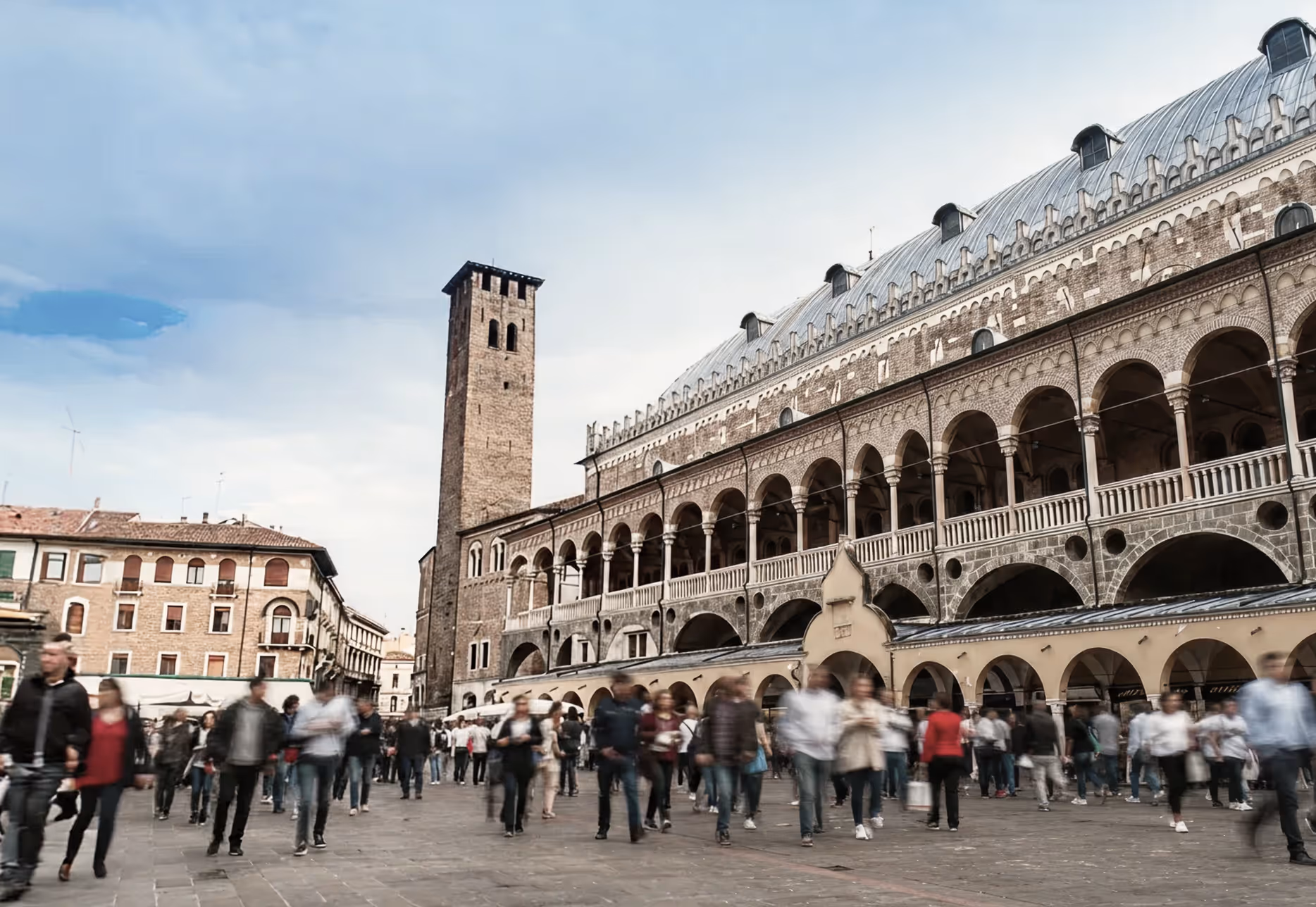 Persone che camminano in una piazza davanti a un edificio storico con archi e una torre in pietra.