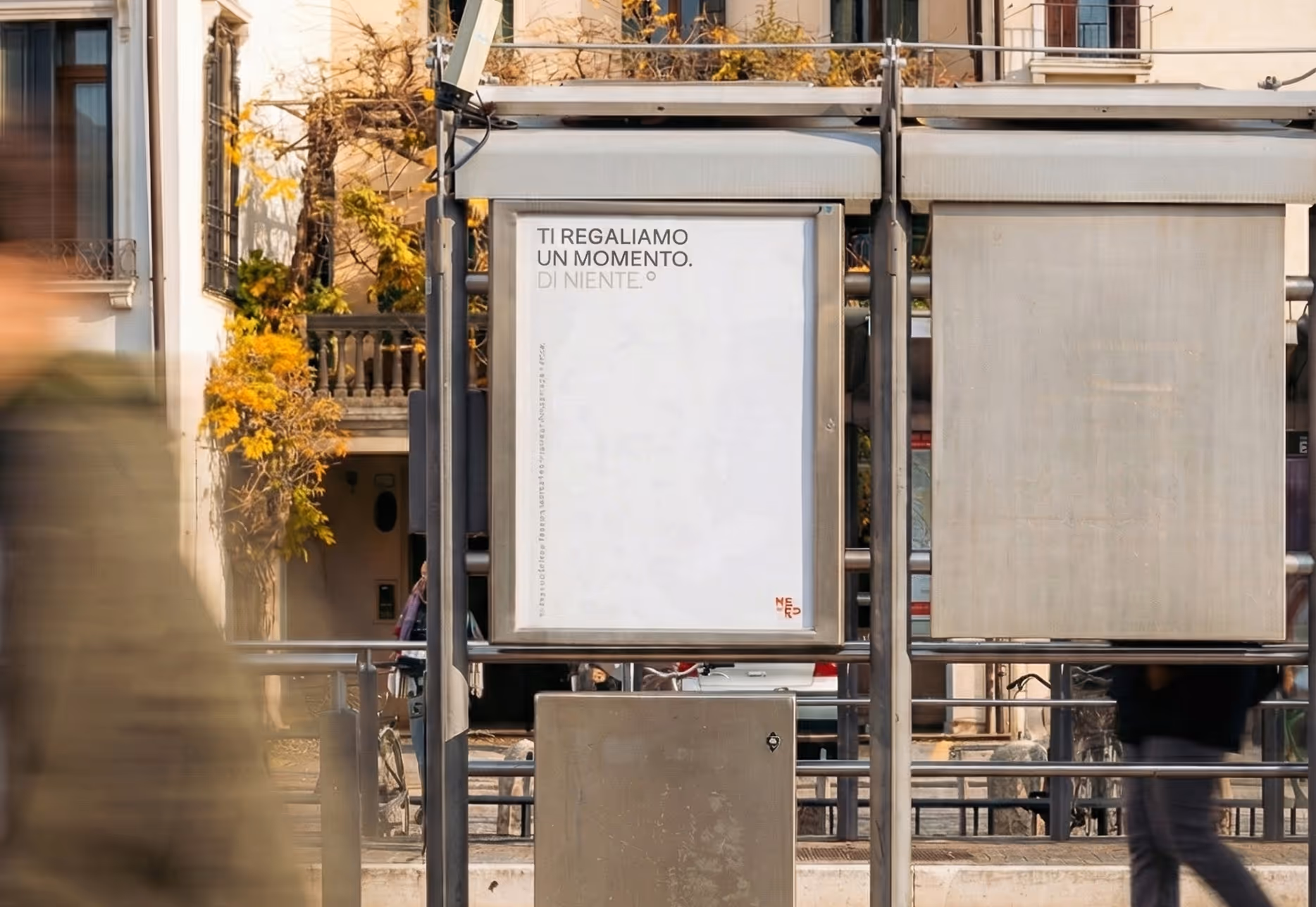 Cartellone pubblicitario vuoto in una fermata dell'autobus in città, con persone sfocate che camminano davanti.