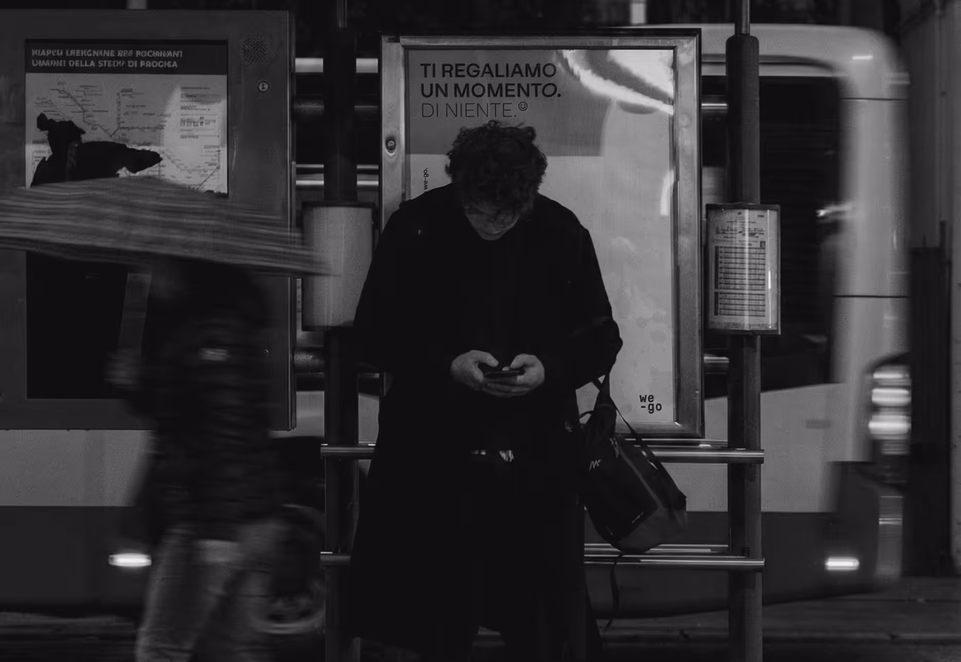 Uomo con giacca nera in piedi a una fermata dell'autobus che guarda il telefono, a sinistra una persona sfocata con un ombrello e a destra un autobus in movimento.