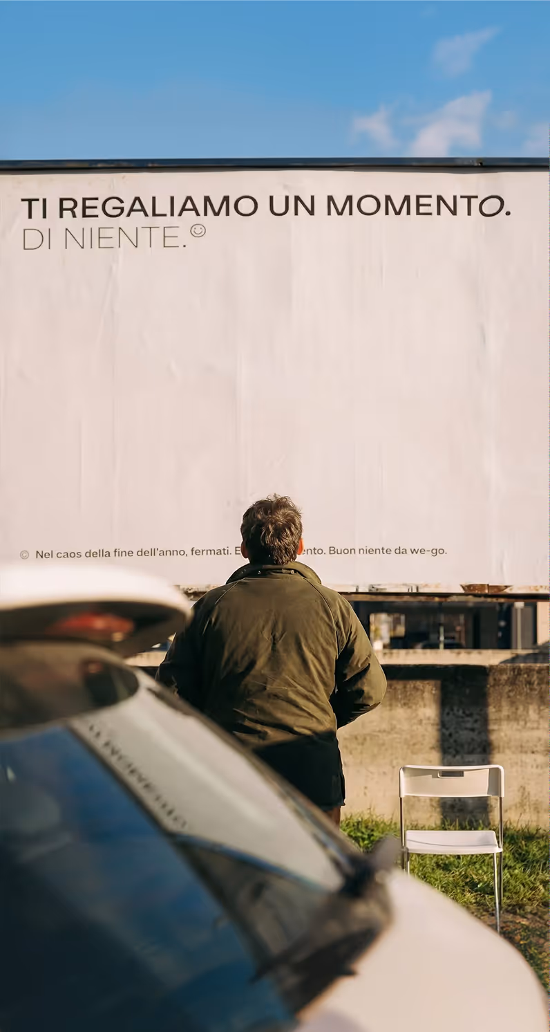Uomo di spalle con giacca verde davanti a un cartellone con scritto "Ti regaliamo un momento. Di niente." e una sedia bianca accanto al muro.